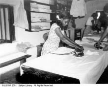 A black and white photograph of a group of people at Beagle Bay doing laundry. There is a young person ironing a large sheet.