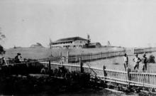 A black and white photograph of Cherbourg Dormitory. There is a long picket fence crossing across the property.