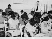 A black and white photograph of a class of young students at Cherbourg Dormitory. They are reading. There is an older adult standing in the back.