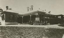 A black and white photograph of Cootamundra Girls Home. A group of residents and staff members are assembled in front of the building, preparing for the photograph.