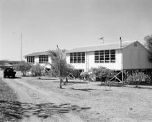 A black and white photograph of two buildings at Croker Island Mission. There is an unsealed road winding to the left of the buildings, and small trees separate the buildings from the road.