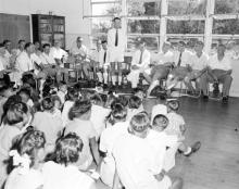 A black and white photograph of people assembled inside a building at Croker Island Mission. The adults are seated, while the children are sitting on the floor. They are all listening to one adult who is standing and speaking.