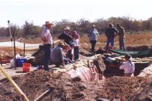 Doctor Judith Field stands in a trench addressing 11 individuals kneeling and standing around the outside.