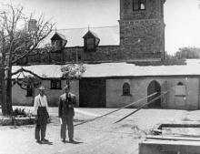 A black and white photograph of the stone-masonry rear of St Francis House. There is a large, wilting tree and two people are standing in front of the tree.