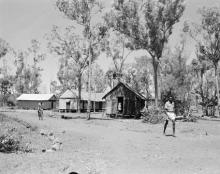 A black and white photograph of Groote Eyelandt Mission. Several small buildings are among tall trees, and a person is riding a bicycle along an unpaved track. 