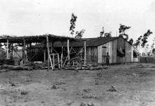 A black and white phoograph of a building at Groote Eyelandt Mission. Beside the building is a shelter roughly constructed from wooden planks, topped with branches as a makeshift roof.