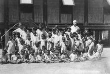 A black and white photograph of a group of children at the Kahlin Compound. The children are assembled, either sitting or standing, in preparation for a photograph. An adult is standing in the back.,