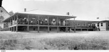 A black and white photograph of the rear porch of Kennion House. There are a large group of people on the porch, either sitting or gazing out across the grass field. 