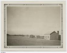 A black and white photograph of several buildings at Koonibba Lutheran Children's Home. The buildings are small, with stone-masonry and coarse plaster walls. Each building has two windows separated by a single door as well as a chimney.