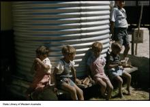 A colour photograph of a few young children at Marribank Mission. They are seated next to a water holding tank constructed from corrugated iron sheet metal.
