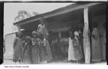 A black and white photograph of a group of people at Moore River Native Settlement. They are standing underneath a shelter made out of corrugated iron.