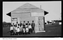 A black and white photograph of a group of people at Moore River Native Settlement. The children are all standing in front of a building which is made out of corrugated iron sheeting. The children are assembled together, with staff standing in the back row, and they are all are preparing for their photograph to be taken.