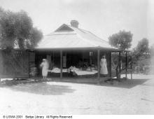 A black and white photograph of a medical ward at Moore River Native Settlement. There are a group of people lying in bed on the porch. There are staff members standing near them.