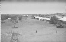 A black and white photograph of Mount Margaret Mission. There are several small buildings scattered among trees, with a large open clearing in front of the buildings. There is a person riding their bicycle in the clearing.