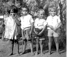 A black and white photograph of young children at Mount Margaret Mission. They are standing side-to-side in a row. The child on the left is hiding a laugh.