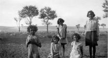 A black and white photograph of a group of children at Mount Margaret Mission. Some children are seated on the grass, and some are standing. They are smiling and looking at the photographer.