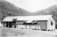 A black and white photograph people at Palm Island Dormitory. They are standing side-to-side in a row. They are outside and in front of a building constructed of corrugated iron sheet metal.