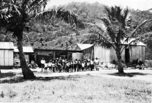 A black and white photograph of people at Palm Island Dormitory. They are standing side-to-side in front of a building constructed of corrugated iron sheet metal. There are two palm trees, one at either side of the group.