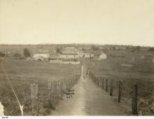 A sepia-toned photograph of Point McLeay Mission. A narrow, unpaved track is leading down to the Mission. The mission consists of several small buildings clustered together in a large, open field.