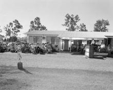 A black and white photograph of a crowd at Retta Dixon Home. They are seated outside in the open are are listening to a person speaking at a podium.