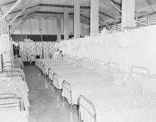 A black and white photograph of the sleeping quarters at Retta Dixon Home. Arranged in rows are a large number of metal framed beds. A single person is fitting sheets over the beds.