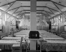 A black and white photograph of the sleeping quarters at Retta Dixon Home. Arranged in rows are a large number of metal framed beds. A single person is seated on one bed.