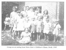 A black and white photograph of a group of children from Sister Kate's Children's Home. They are assembled together and preparing to be photographed. They are standing on the grass outside of a brick-faced building.