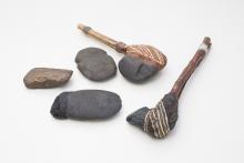 Two ancient tools comprised of a painted stone head afixed to a wooden handle rest alongside three stone axe-head artefacts.