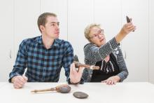 A man holds an ancient stone hatchet while the woman next to him examines a stone axe-head. More examples of ancient tools rest on the table infront of them.