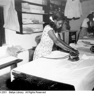 A black and white photograph of a group of people at Beagle Bay doing laundry. There is a young person ironing a large sheet.