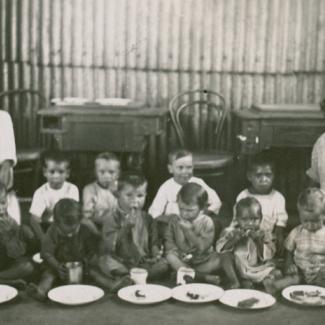 A black and white photograph of a group of young children at The Bungalow. They are eating and drinking while sitting on the floor. There are two adults seated to the left and right of this group.