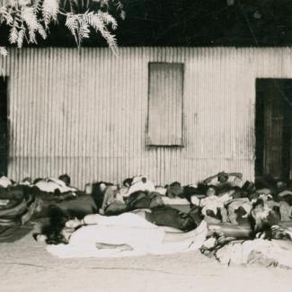 A black and white photograph of a large group of people at The Bungalow. They are sleeping on the ground outside a building. They are lying on blankets, and three or four people share one blanket.