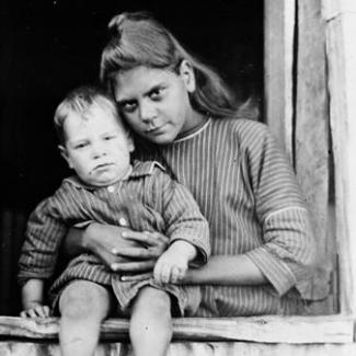 A black and white photograph of a child and adult at The Bungalow. The adult is standing near a window and is holding a toddler who is sitting on the window-sill.