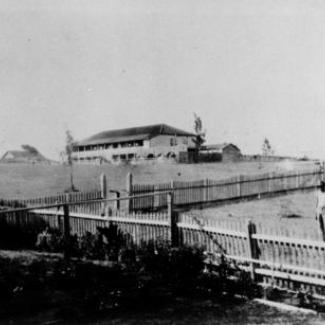 A black and white photograph of Cherbourg Dormitory. There is a long picket fence crossing across the property.