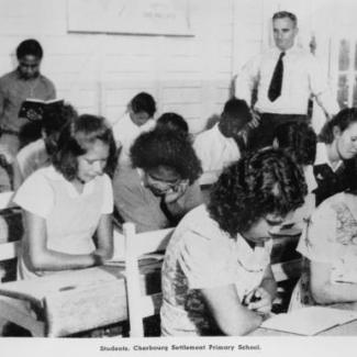 A black and white photograph of a class of young students at Cherbourg Dormitory. They are reading. There is an older adult standing in the back.