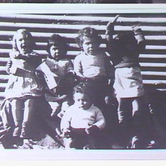A black and white photograph of a small group of young children from Colebrook Home. They are sitting beside a corrugated iron sheet and are singing.