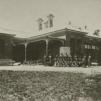 A black and white photograph of Cootamundra Girls Home. A group of residents and staff members are assembled in front of the building, preparing for the photograph.