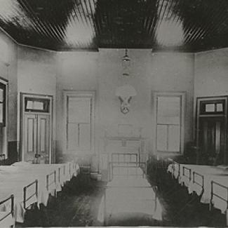 A black and white photograph of the sleeping quarters of Cootamundra Girls' Home. Beds line the left, right and centre of this room.
