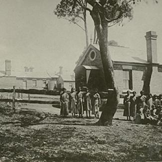 A black and white photograph of a building at Cootamundra Girls' Home. Standing in front of the building there are a group of  residents and  staff members.