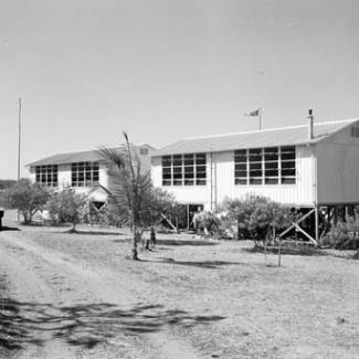 A black and white photograph of two buildings at Croker Island Mission. There is an unsealed road winding to the left of the buildings, and small trees separate the buildings from the road.
