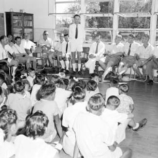 A black and white photograph of people assembled inside a building at Croker Island Mission. The adults are seated, while the children are sitting on the floor. They are all listening to one adult who is standing and speaking.