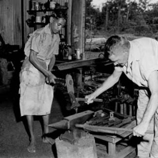 A black and white photograph of the forge at Garden Point Mission. An older child and an adult are using hammers at an anvil.