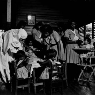 A black and white photograph of young children at Garden Point Mission. They are sitting at a table, eating and drinking, and are being assisted by several adults.