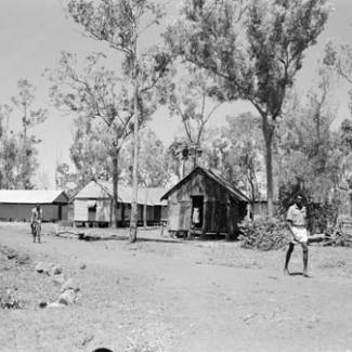 A black and white photograph of Groote Eyelandt Mission. Several small buildings are among tall trees, and a person is riding a bicycle along an unpaved track. 