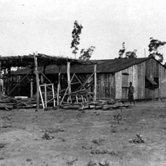 A black and white phoograph of a building at Groote Eyelandt Mission. Beside the building is a shelter roughly constructed from wooden planks, topped with branches as a makeshift roof.