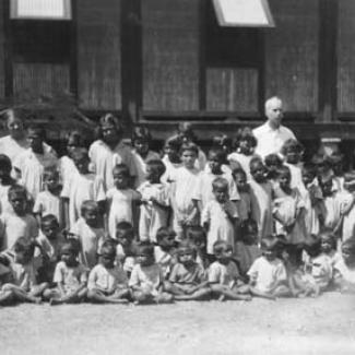 A black and white photograph of a group of children at the Kahlin Compound. The children are assembled, either sitting or standing, in preparation for a photograph. An adult is standing in the back.,