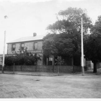 A black and white photograph of the front of Kennion House. The building has a brick face, and several trees which shield the ground floor windows from the street.