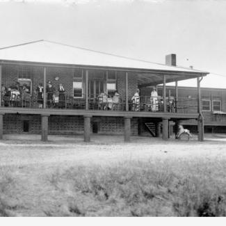 A black and white photograph of the rear porch of Kennion House. There are a large group of people on the porch, either sitting or gazing out across the grass field. 