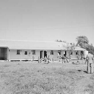 A black and white photograph of a building at Kinchela Boys' home. A group of children are running in front of the building. An adult is watching them.