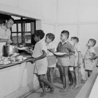 A black and white photograph of a group of children at Kinchela Boys' Home. They are are queued in the cafeteria, waiting to be served a meal by a staff member.